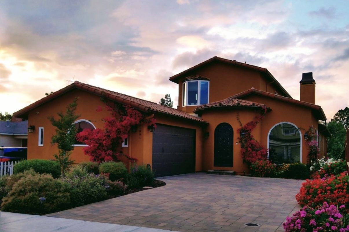 Original exterior photo of a two-story stucco home with a tile roof, a garage and a paver driveway, at dusk