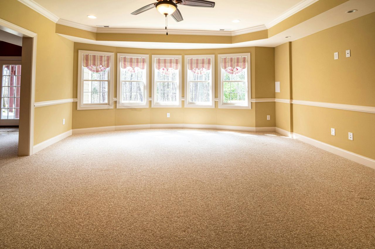 Original photo of an empty living room with yellow walls, beige carpet, a ceiling fan and a row of five windows